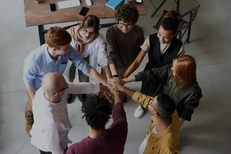 An overhead photo of a diverse team of 8 people standing in a circle with their hands in the middle joined - Include with Dr. Liz Wilson - diversity and inclusion expert