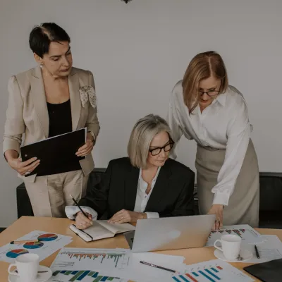 This is a photo of a three women at a table in a meeting room referring to a laptop and printed documents working on an inclusion strategy with Dr. Liz Wilson from Include Inc. - Diversity and inclusion expert consultant